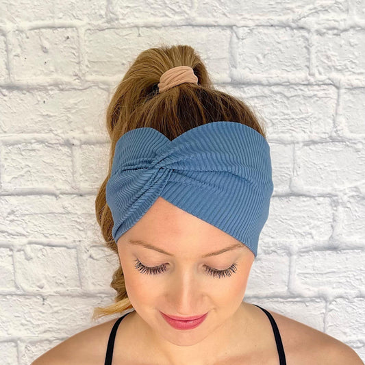 Woman with hair in curled ponytail wearing wide, twisted, stretchy headband in blue ribbed fabric.