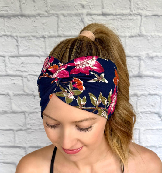 Woman with hair in ponytail wearing wide, twisted, navy color headband with pink, orange, and green floral print.