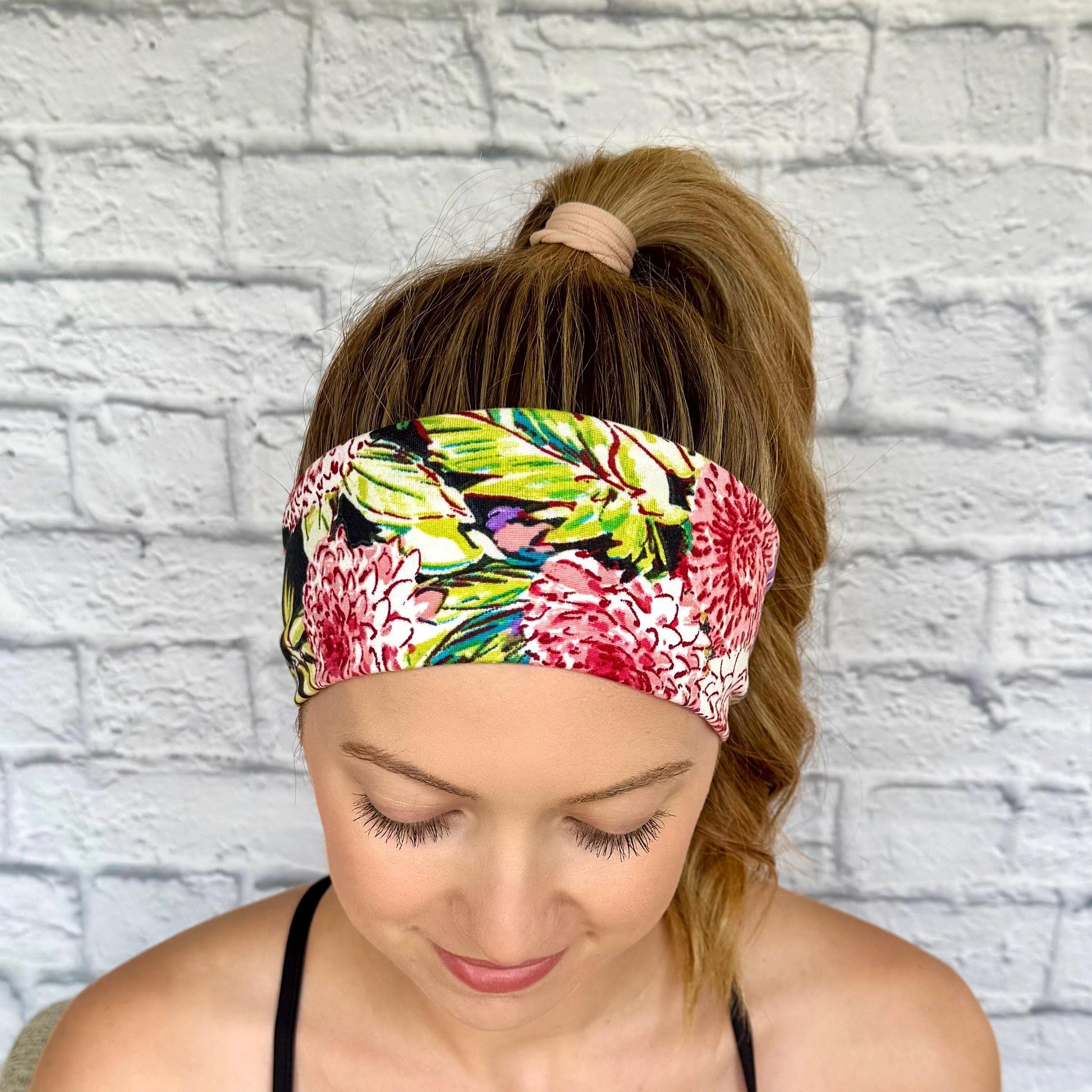 Woman with hair in ponytail wearing flat, stretchy headband in black with lime green, pink, and white floral print.