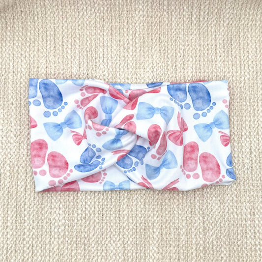 Wide, twisted, white headband with pink and blue baby feet and bow print.