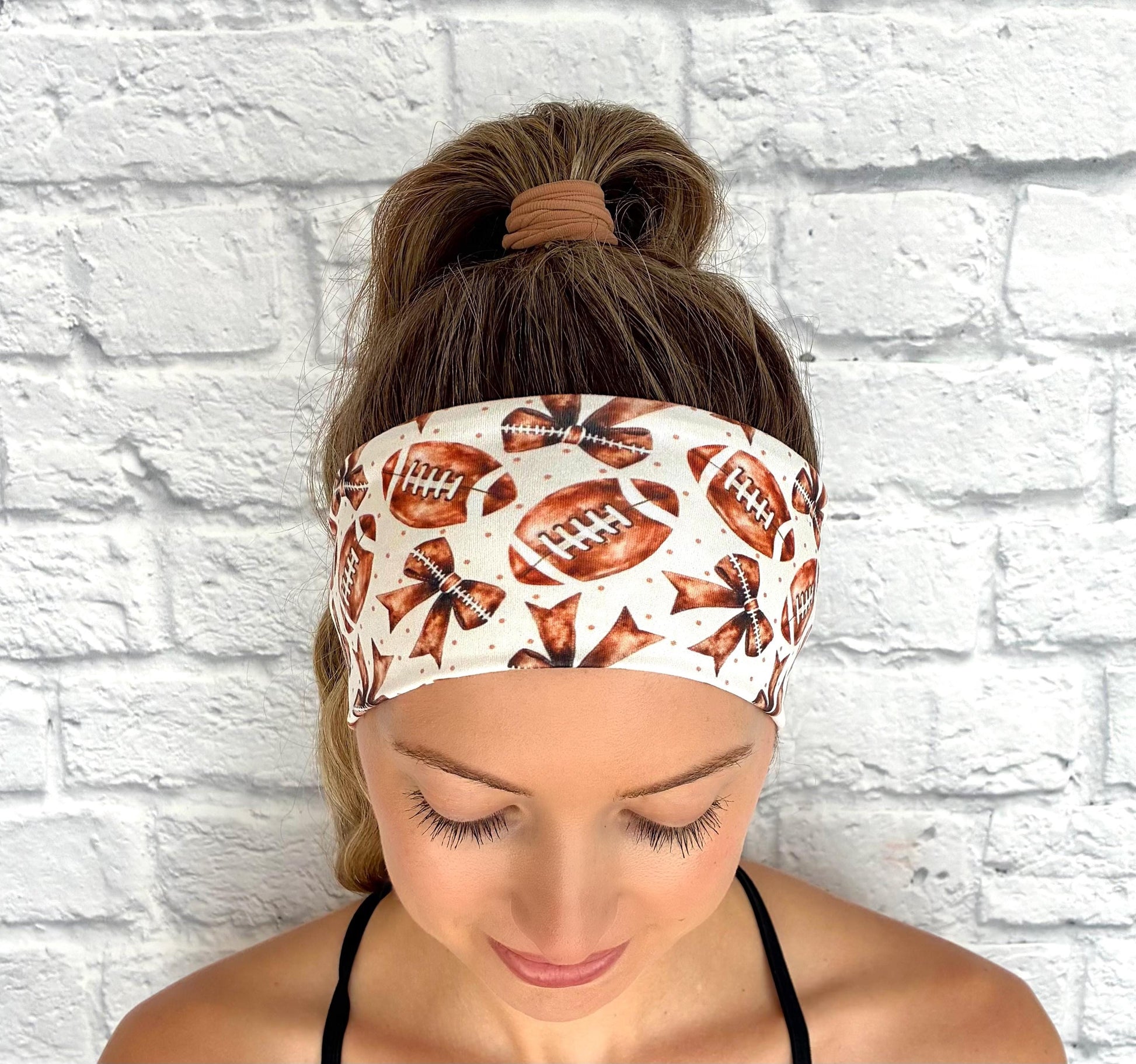 Woman with hair in ponytail wearing flat, stretchy headband in white with brown football and bow print.