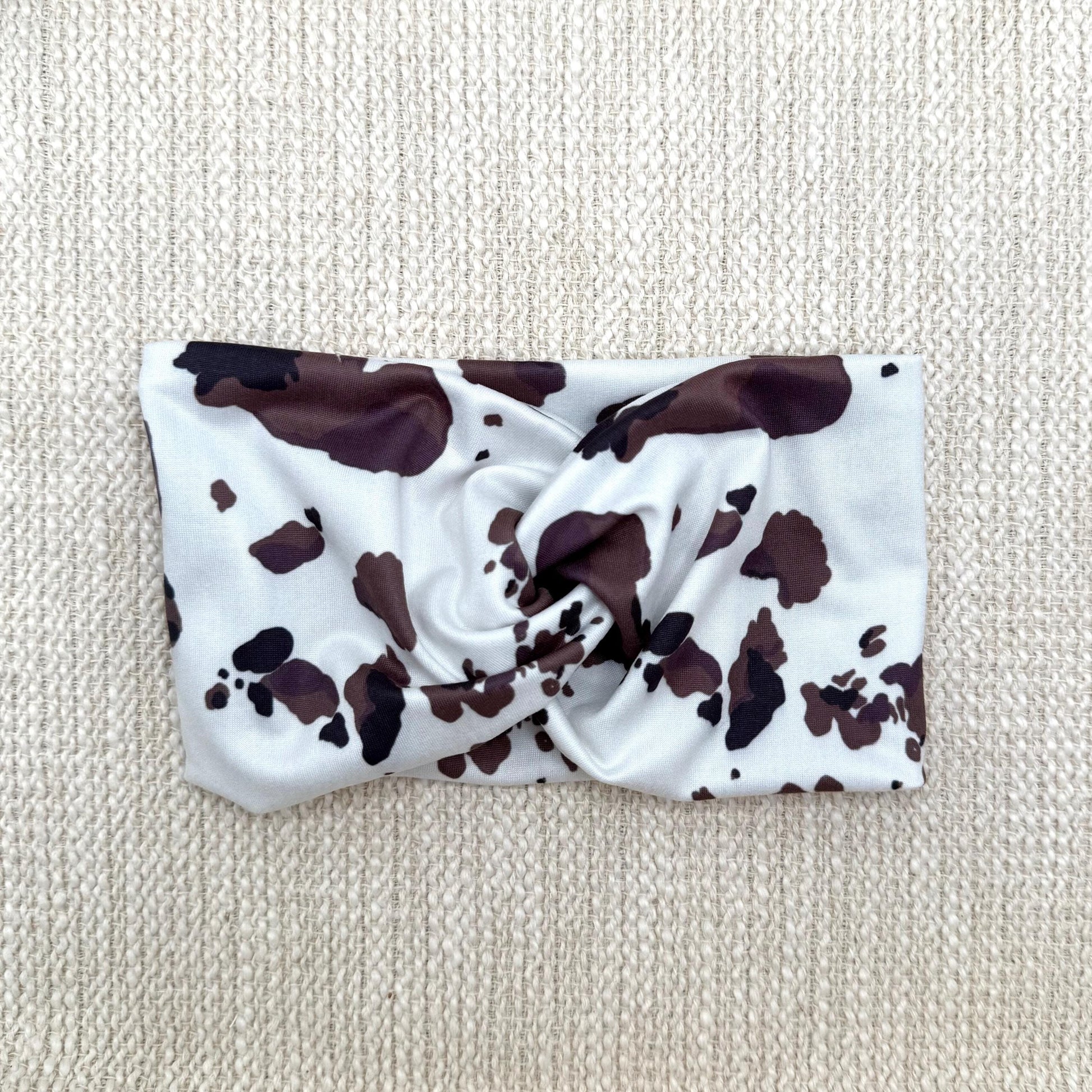 Wide, twisted headband with cow print displayed on a cream color background.