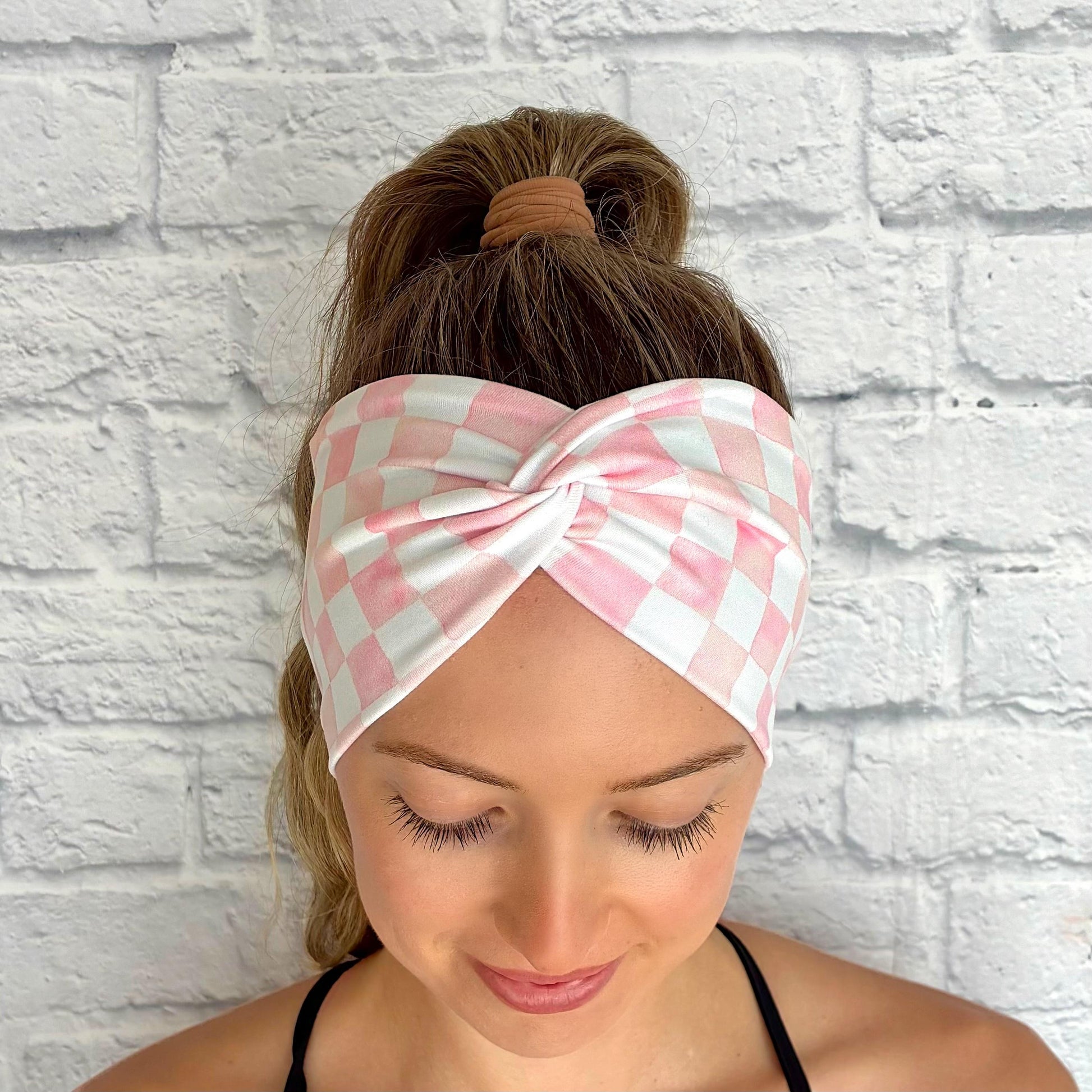 Woman with hair in ponytail wearing a wide, twisted headband in light pink and white checker print.
