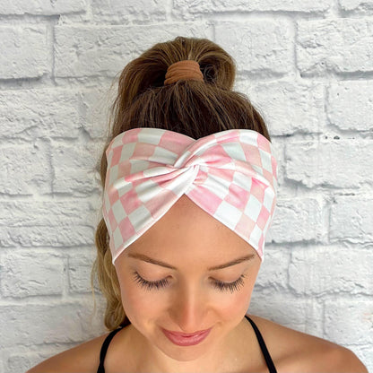 Woman with hair in ponytail wearing a wide, twisted headband in light pink and white checker print.