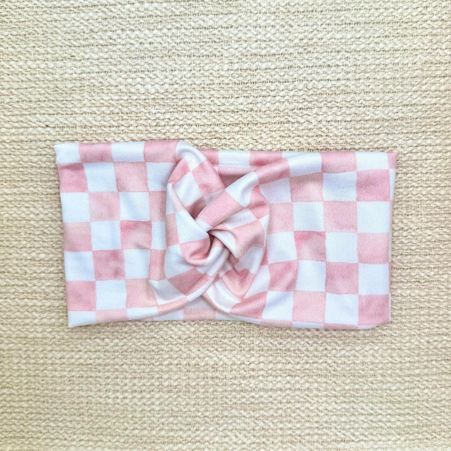 Woman with hair in ponytail wearing a wide, twisted headband in light pink and white checker print.