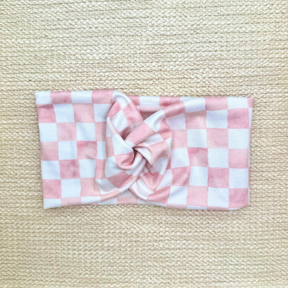 Woman with hair in ponytail wearing a wide, twisted headband in light pink and white checker print.