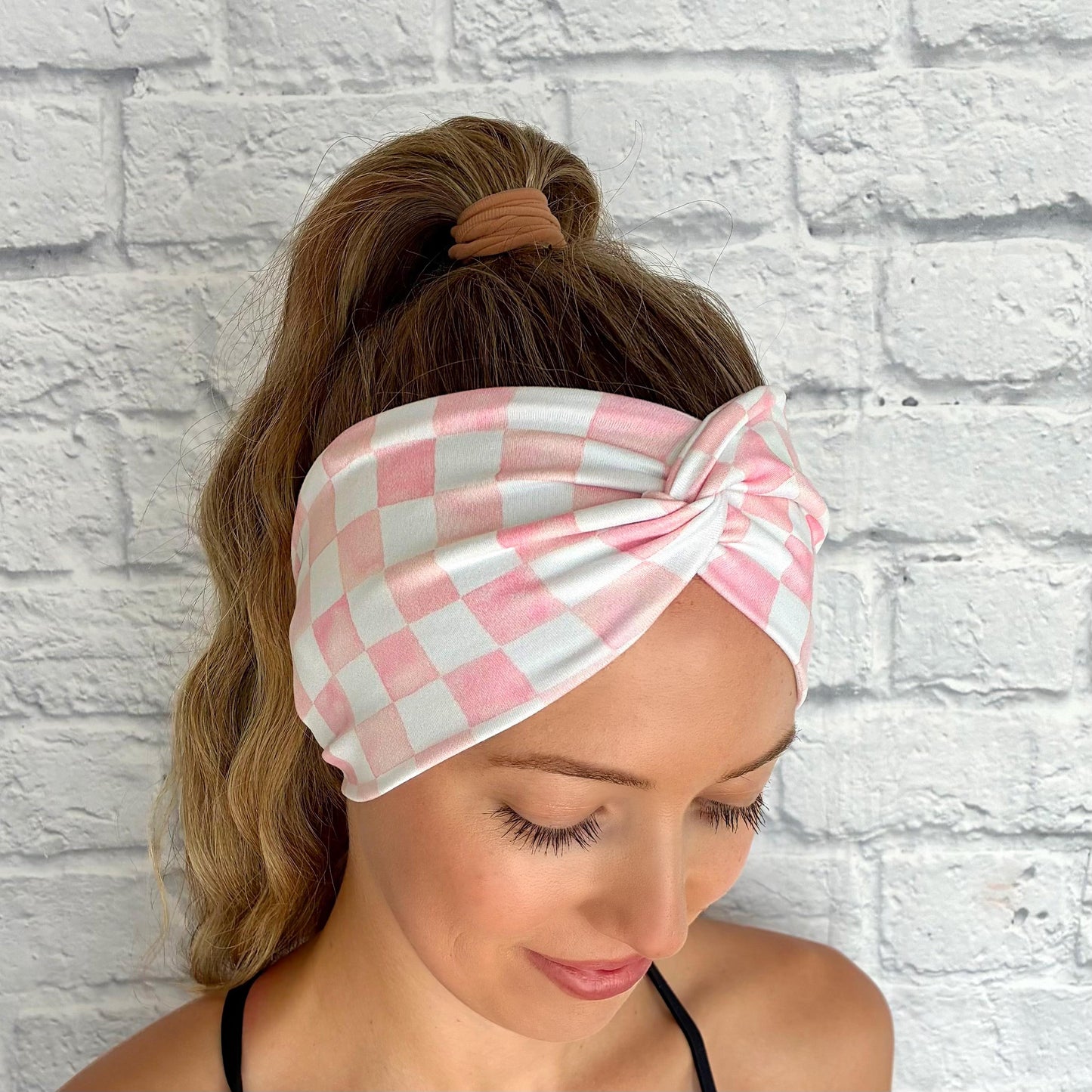Woman with hair in ponytail wearing a wide, twisted headband in light pink and white checker print.