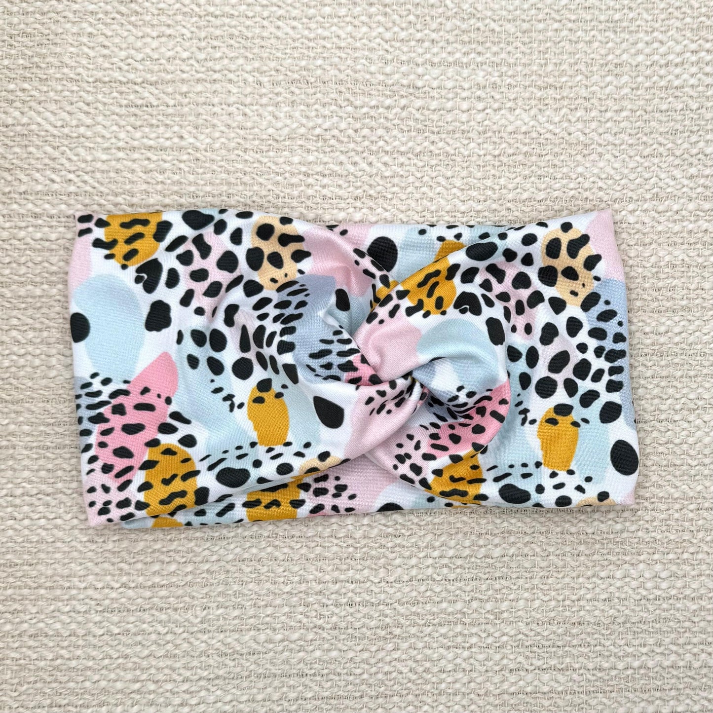 Wide, twisted headband in white with pink, mustard yellow, and blue splotchy pattern print with black leopard print.