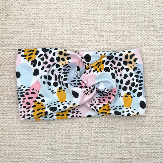 Wide, twisted headband in white with pink, mustard yellow, and blue splotchy pattern print with black leopard print.