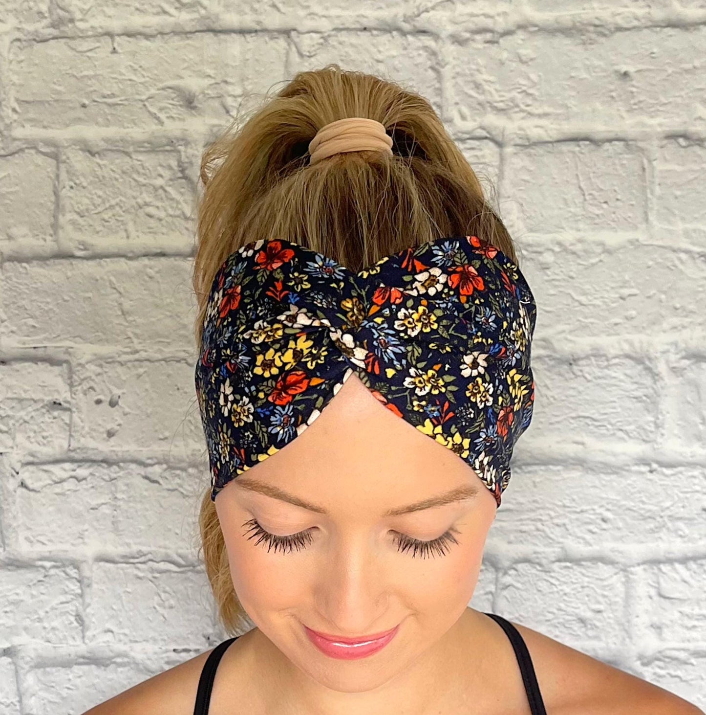 Woman with hair in ponytail wearing wide, twisted headband in navy with red, white, yellow, and blue small flower print.