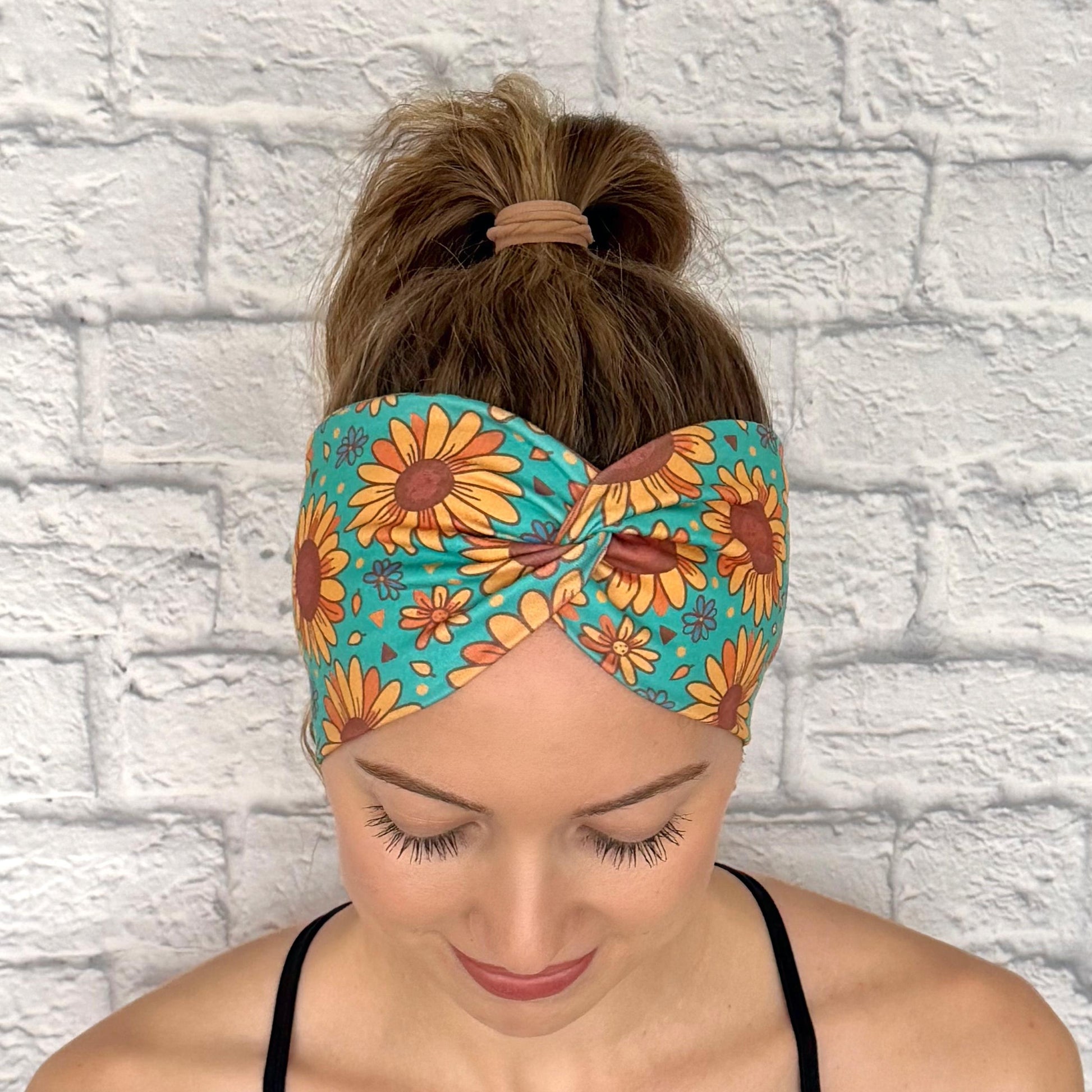 Woman with hair in ponytail wearing wide, twisted headband in teal color with sunflower print.