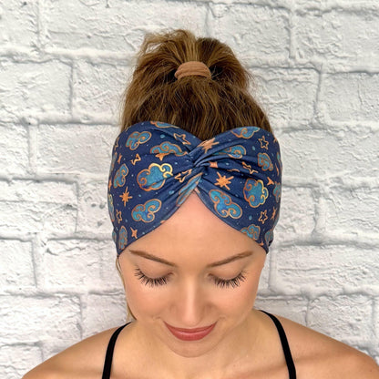 Woman with hair in ponytail wearing wide, twisted headband in navy blue with clouds, stars, and space theme print.