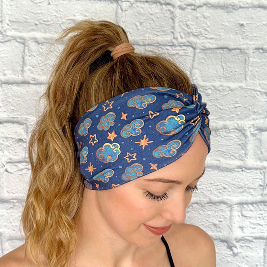 Woman with hair in ponytail wearing wide, twisted headband in navy blue with clouds, stars, and space theme print.