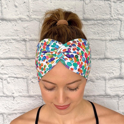 Woman with hair in ponytail wearing wide, twisted headband in white with rainbow color polka dot print.