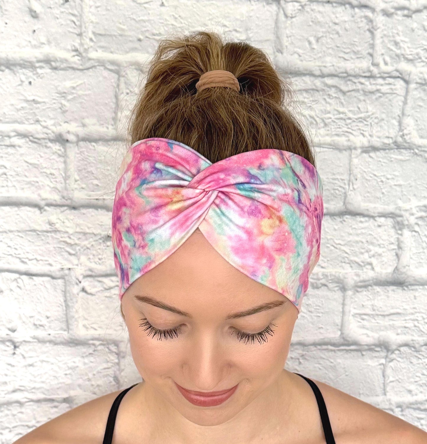 Woman with hair in ponytail wearing wide, twisted headband with pink and multicolor tie dye print.
