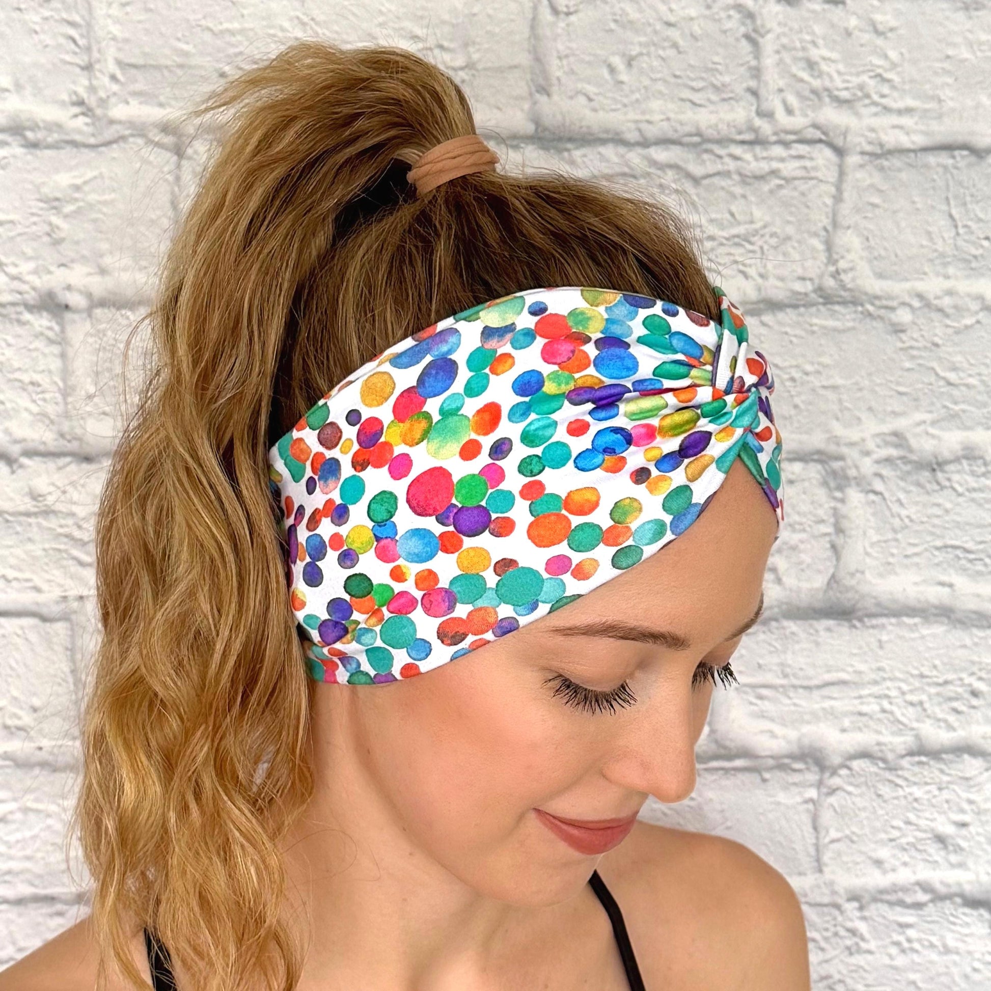 Woman with hair in ponytail wearing wide, twisted headband in white with rainbow color polka dot print.