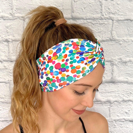 Woman with hair in ponytail wearing wide, twisted headband in white with rainbow color polka dot print.