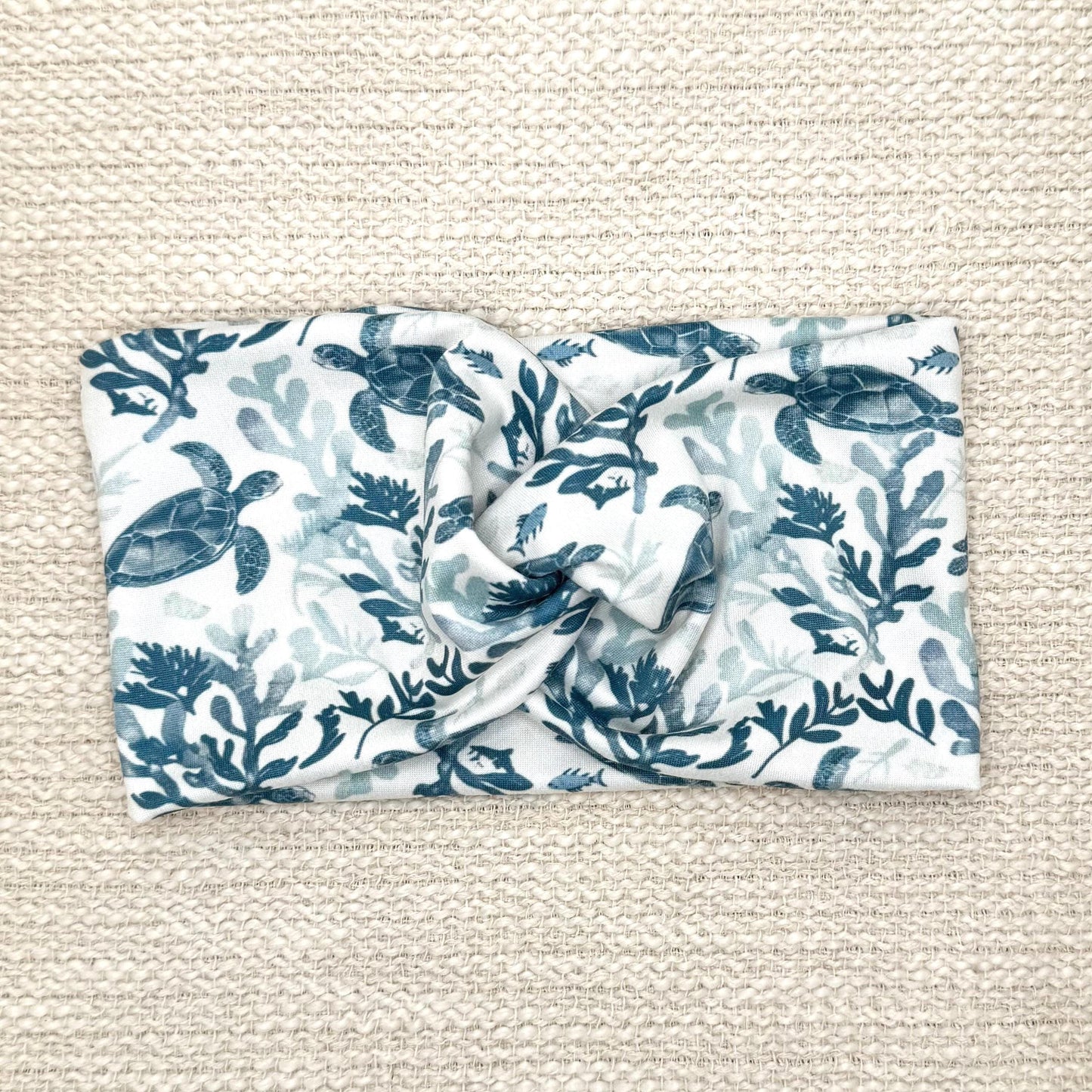 Wide, twisted, white headband with sea turtle print, displayed on cream background.