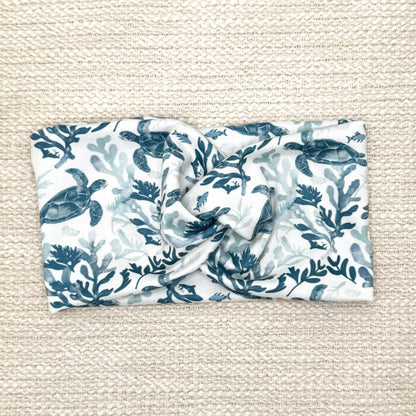 Wide, twisted, white headband with sea turtle print, displayed on cream background.