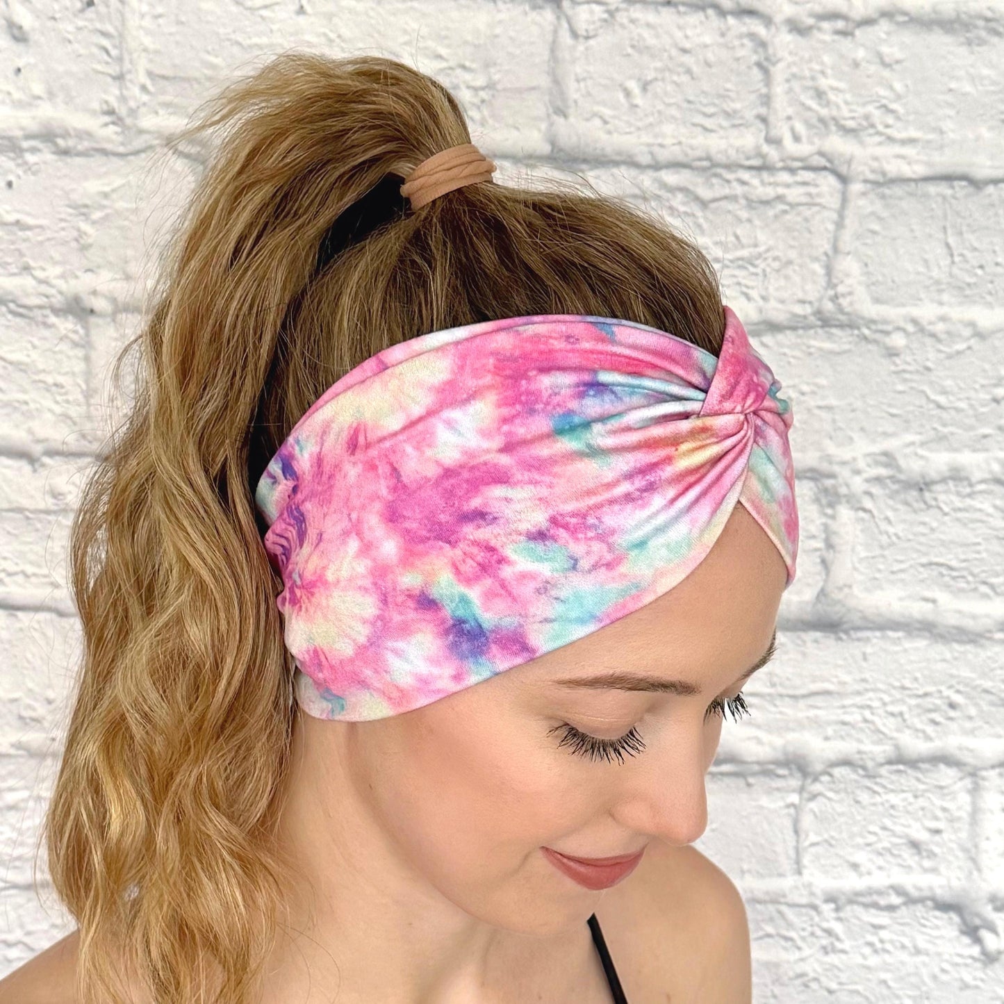Woman with hair in ponytail wearing wide, twisted headband with pink and multicolor tie dye print.