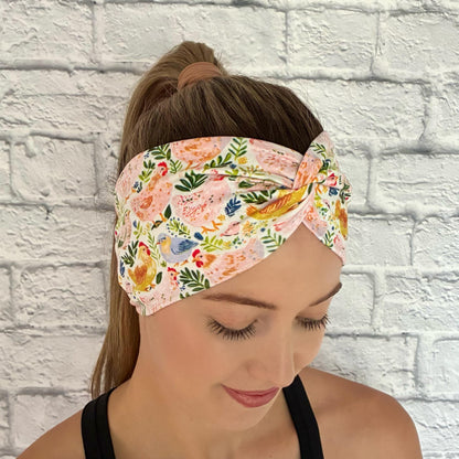 Woman with hair in a ponytail wearing a wide, twisted headband with a colorful chicken and floral print.