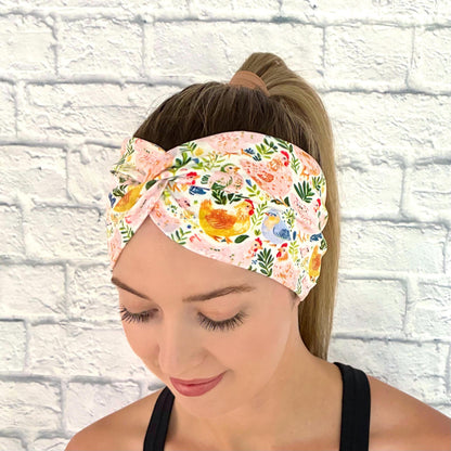 Woman with hair in a ponytail wearing a wide, twisted headband with a colorful chicken and floral print.