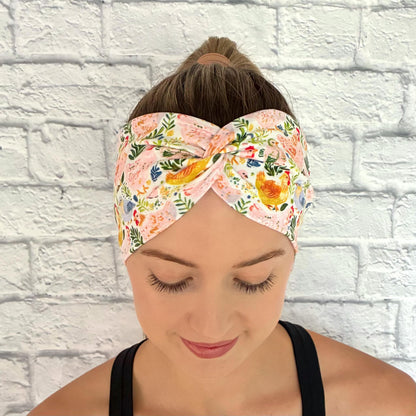 Woman with hair in a ponytail wearing a wide, twisted headband with a colorful chicken and floral print.