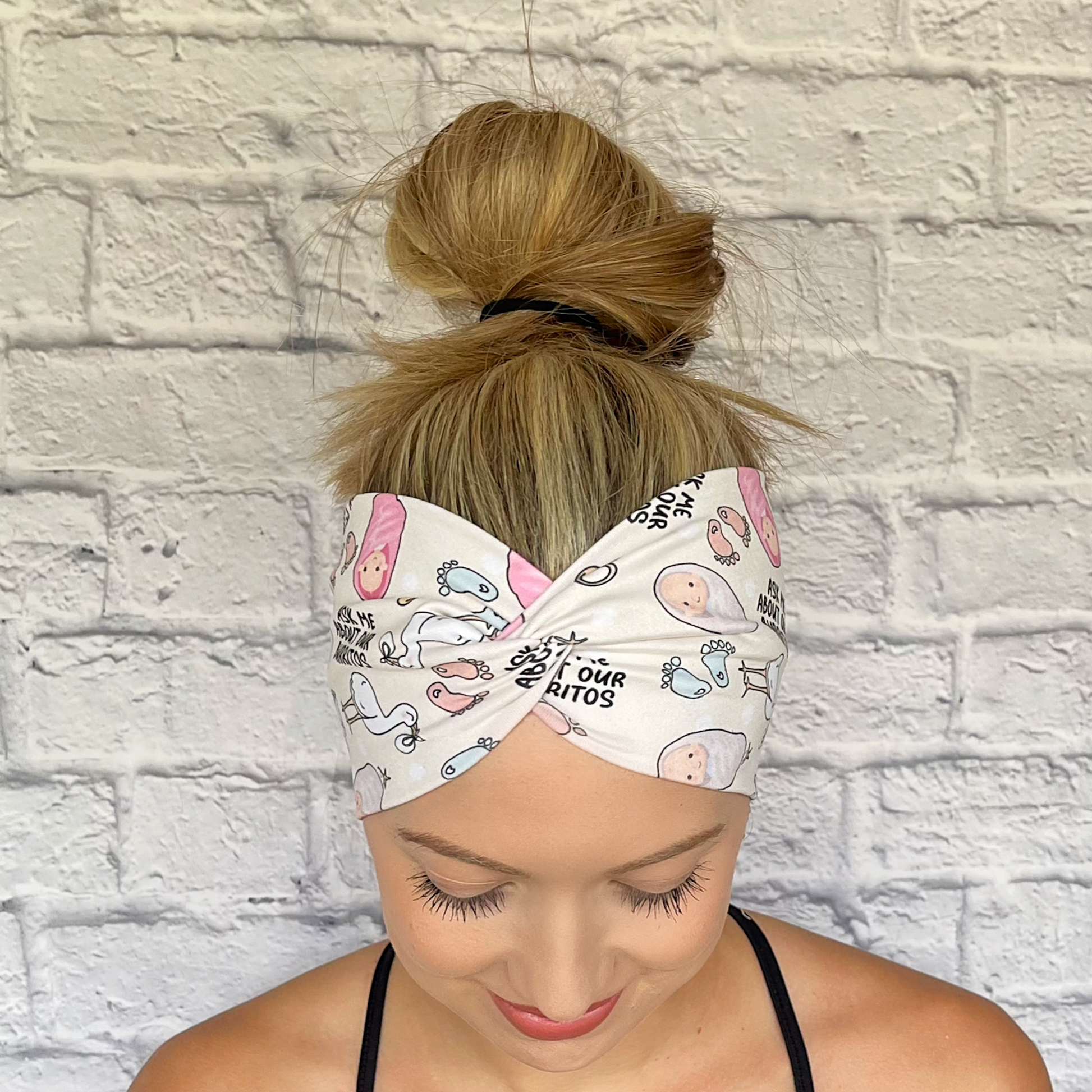 Woman with hair in bun wearing wide twisted headband. The headband is cream color with baby theme print: swaddled babies, baby feet, storks, pacifiers, and "Ask Me About Our Burritos" print throughout.