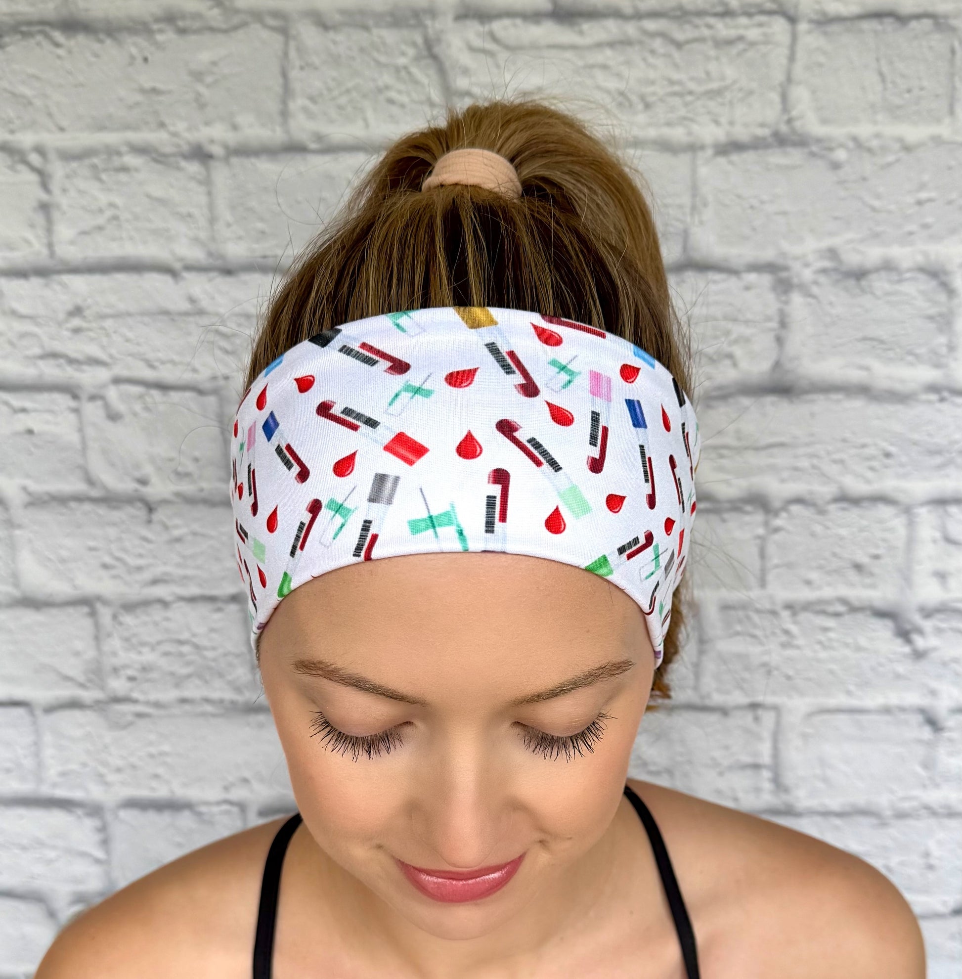 Woman with hair in ponytail wearing white headband with phlebotomy theme print- blood vials, blood drops, and syringes.