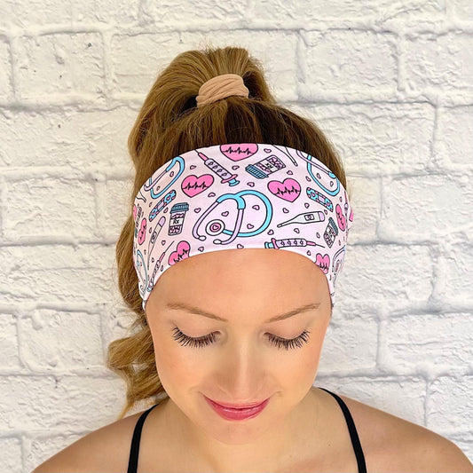 Woman with hair in curled ponytail wearing flat, stretchy headband in light pink with pink, purple, and blue medical theme print.