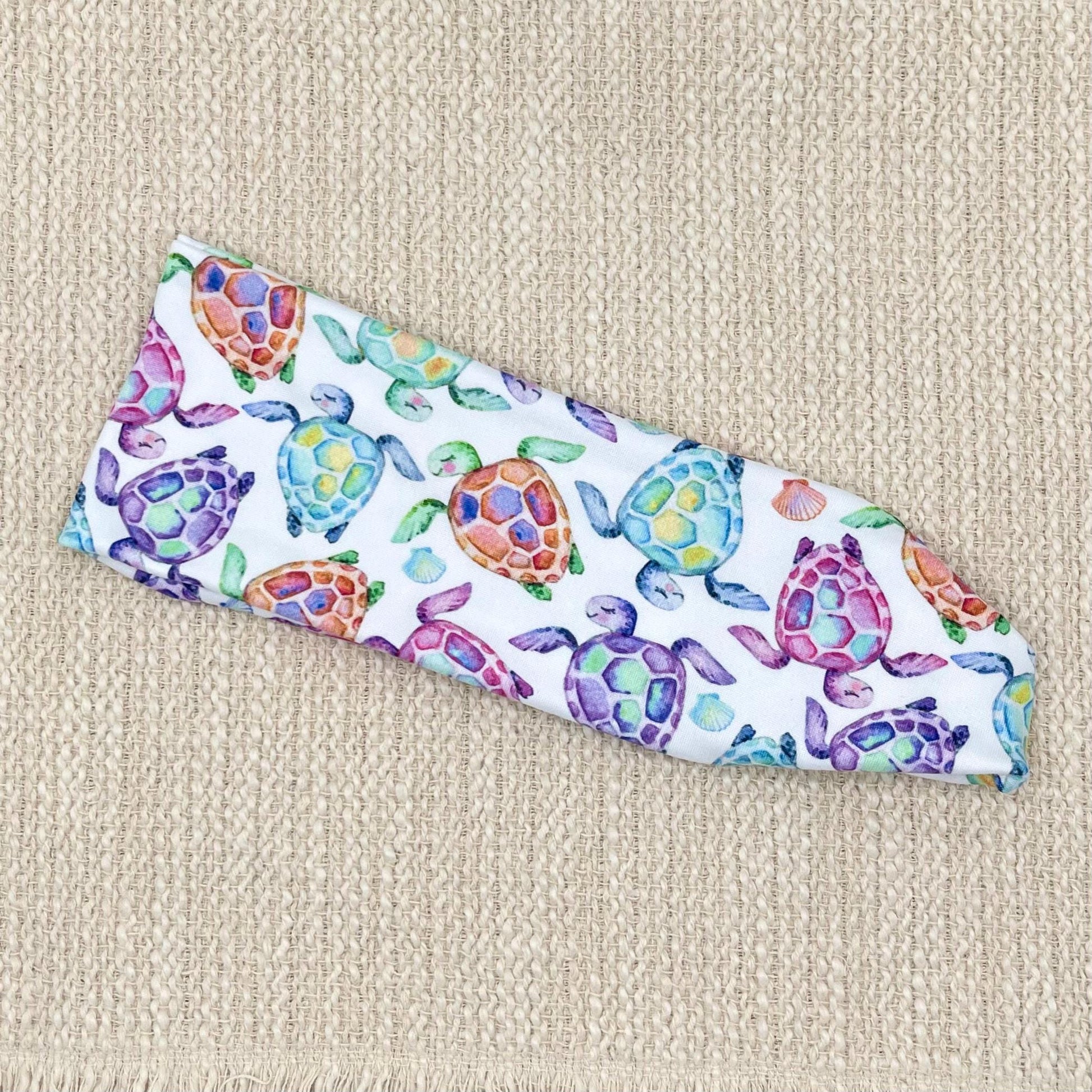 Flat, stretchy, white headband with sea turtle print, displayed on a cream background.